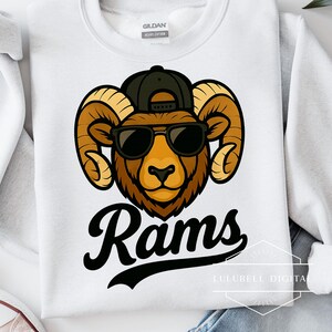 May include: White sweatshirt featuring a cartoon ram wearing a black cap and sunglasses. The ram has brown fur and large curved horns. The word "Rams" is written in black script below the graphic.