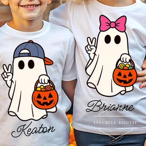 May include: White t-shirts with Halloween ghost designs. One ghost wears a blue and orange baseball cap and holds a jack-o'-lantern candy bucket. The other ghost has a pink bow and also holds a jack-o'-lantern candy bucket. The names "Keaton" and "Brianne" are below the ghosts.