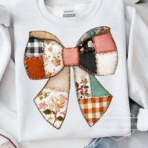 May include: White sweatshirt featuring a large, patchwork bow design. The bow is composed of various fabric patterns, including floral, plaid, and solid colors, stitched together with visible seams. The sweatshirt has a crew neck and long sleeves.