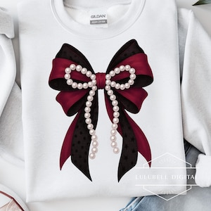 Op de afbeelding: Witte sweatshirt met een groot strikdesign. De strik heeft zwarte en bordeauxrode linten, versierd met een parelsnoer. Het zwarte lint heeft een patroon van kleine stippen en sterren.