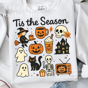 May include: White sweatshirt with a colorful Halloween-themed graphic. The design includes a witch on a broom, pumpkins, ghosts, a black cat, a skull, and the text "Tis the Season".