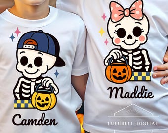 Op de afbeelding: Witte T-shirts met Halloween-skeletontwerpen. Eén skelet draagt een blauwe baseballpet en de naam "Camden" staat eronder. Het andere skelet heeft een roze strik en de naam "Maddie". Elk houdt een pompoen vast.