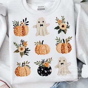 Op de afbeelding: Witte sweatshirt met een herhalend patroon van pompoenen, bloemstukken en geesten. De pompoenen zijn oranje en bruin met groene stelen. De geesten hebben strikjes. De sweatshirt is van zwaar katoen.