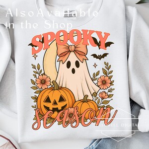 May include: White sweatshirt with a retro-style Halloween design. The design features a ghost with a bow, pumpkins, flowers, and the words "Spooky Season" in orange. A bat and stars complete the design.