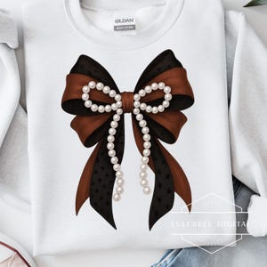 May include: White sweatshirt featuring a large bow design. The bow has layers of brown and black ribbons, with a pearl strand in the center and hanging down. The black ribbon has a star pattern. The brand tag is visible.