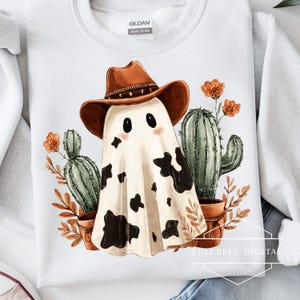 May include: White sweatshirt featuring a cartoon ghost wearing a brown cowboy hat with black spots. The design includes cacti, orange flowers, and brown leaves in terracotta pots. The brand name "Gildan" is visible.