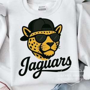 May include: White sweatshirt featuring a cartoon jaguar head wearing sunglasses and a black cap. The word "Jaguars" is written in a stylish script below the jaguar graphic. The sweatshirt is a Gildan Heavy Cotton.