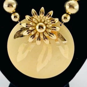 May include: Gold-toned necklace featuring a large circular pendant with a floral design and a smaller flower at the center. The necklace has a chain with round beads and a herringbone chain. The jewelry is displayed on a black background.