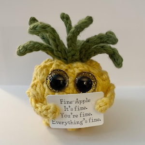 Op de afbeelding: Een gehaakte ananas met grote, glinsterende ogen en groene bladeren. De gele vrucht houdt een klein bordje vast met de tekst "Fine-Apple, It's fine. You're fine. Everything's fine."