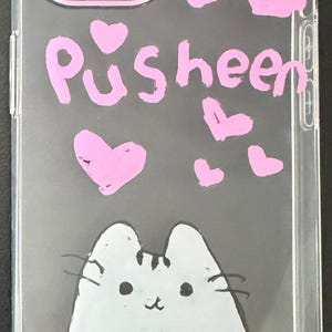 Puede incluir: Funda transparente para tel&eacute;fono con una ilustraci&oacute;n de gato de dibujos animados y la palabra "Pusheen" en rosa. Corazones rosas est&aacute;n dispersos por la funda. La funda tiene un parachoques protector en los bordes.