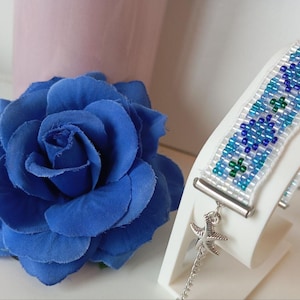 May include: A beaded bracelet with a white base, featuring blue and green floral beadwork. A silver starfish charm dangles from the bracelet. A large blue rose is positioned nearby.