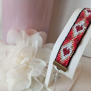 May include: A woven bracelet featuring a heart design in red, white, and gold beads. The bracelet is displayed on a white stand, with a small heart charm hanging from it. The background includes a pink vase and a white flower.