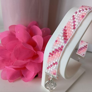 May include: A beaded bracelet and matching earring set on a white display stand. The bracelet and earrings have a pattern of pink, white, and red beads. A silver charm with a tree design hangs from the bracelet. A pink flower and vase are in the background.