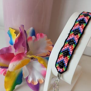 May include: A colourful beaded bracelet with a black border, displaying a geometric pattern in pink, blue, green, and orange. The bracelet is on a white stand, with a vibrant artificial flower in the background.