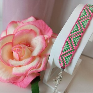 May include: A colourful woven bracelet in pink and green, displayed on a white stand. The bracelet features a repeating pattern of pink and green squares. A large pink and white rose is in the background.