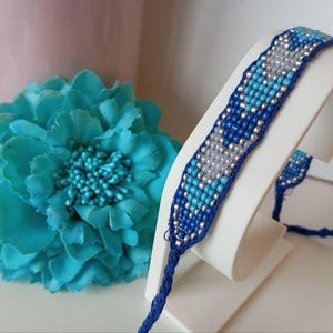 May include: A beaded bracelet in shades of blue, silver, and white is displayed on a white stand. The bracelet features a woven pattern. A large, turquoise fabric flower and a pink vase are in the background.