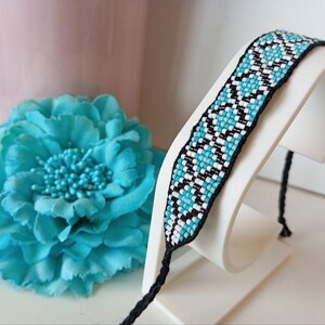 May include: A beaded bracelet with a geometric pattern in turquoise, black, and white is displayed on a white stand. The bracelet has black ties on each end. A large turquoise flower is in the background.