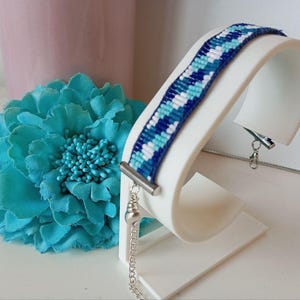 May include: A blue and white woven bracelet on a white display stand. The bracelet has a geometric pattern in dark blue, light blue, and white. A large turquoise fabric flower is in the background.