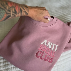 May include: A dusty rose sweatshirt with the embroidered text "ANTI SOCIAL CLUB" in white and pink. The crew neck sweatshirt is folded on a white bedspread.