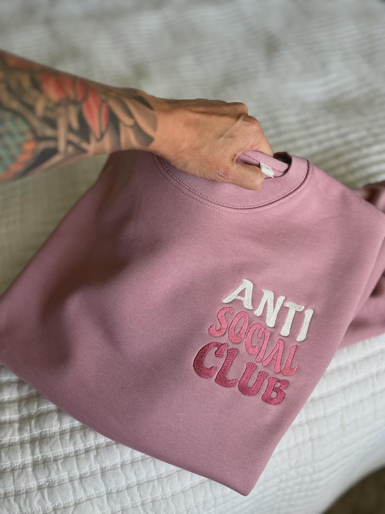 May include: A dusty rose sweatshirt with the embroidered text "ANTI SOCIAL CLUB" in white and pink. The crew neck sweatshirt is folded on a white bedspread.