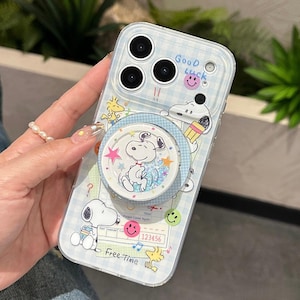 Puede incluir: Funda de teléfono azul claro con diseño de Snoopy. La funda presenta un patrón de cuadros y ilustraciones de Snoopy y Woodstock. Se adjunta un soporte para teléfono a juego con Snoopy. El texto "Good Luck" y "Free Time" son visibles.