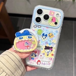 May include: Clear phone case with a blue border and colorful cartoon character stickers. A matching pop socket with a blue character design is also visible. The case and pop socket feature Japanese text and cartoon graphics.