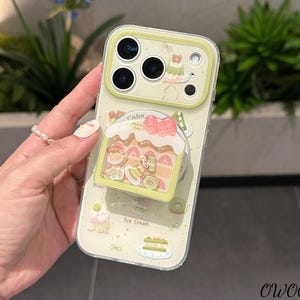 Puede incluir: Una funda transparente para teléfono con un borde verde claro y un diseño de pastel caprichoso. La funda presenta un agarre emergente con una rebanada de pastel, fresas y kiwis. Las palabras "Cake" e "Ice Cream" son visibles.