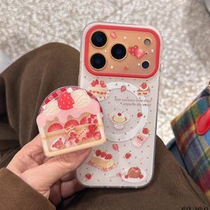 May include: A clear phone case with a strawberry and cake design. The case features a red border and a pop-up cake holder. The design includes illustrations of cakes, strawberries, and the text "Your existence is my brief utopia in this world."