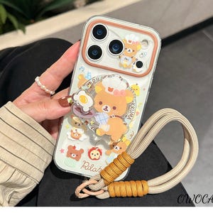 May include: A clear phone case with a brown border featuring a cartoon bear design. The case has a pop socket with a bear and food graphic. A beige and orange lanyard is attached to the case. The text "Rilak" and "OWOCASE" are visible.