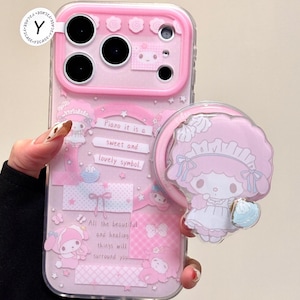 以下が含まれることがあります： マイメロディのデザインとマッチしたポップソケットが付いたピンク色の携帯電話ケース。ケースは透明な縁取りで、「Piano it is a sweet and lovely symbol」というテキストが含まれています。デザインには星、リボン、その他の装飾要素が含まれています。