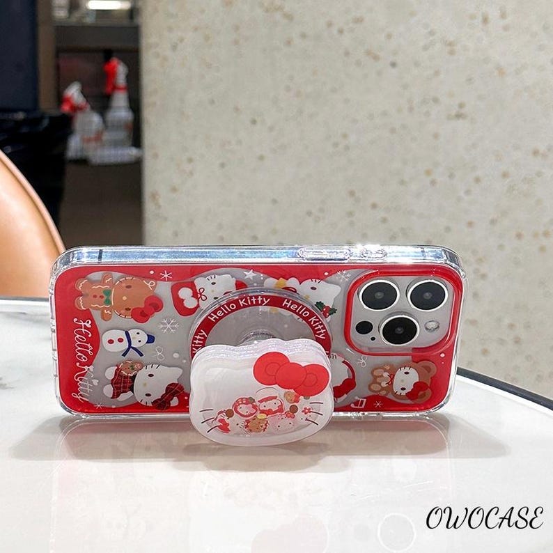 May include: Clear phone case with a red border and a Hello Kitty design. The case features a pop-up grip with a red bow and the words "Hello Kitty". The case has a festive winter theme with gingerbread men and snowmen.