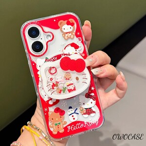 May include: Red and clear phone case featuring Hello Kitty and other festive characters. The case has a pop-up grip with a large red bow and various holiday-themed designs, including a gingerbread man and snowman. The text "Hello Kitty" is visible.