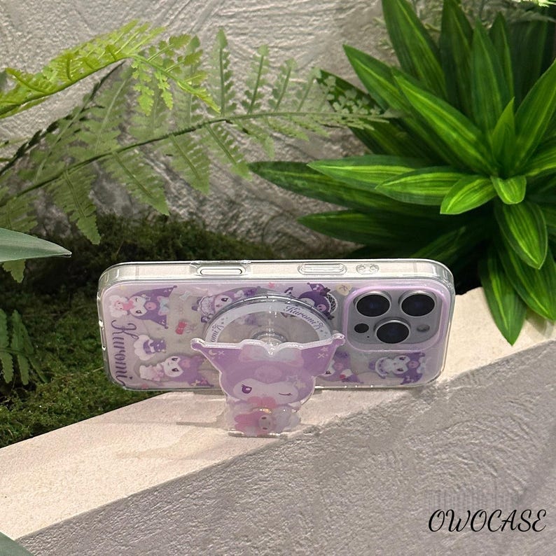 Puede incluir: Funda transparente para tel&eacute;fono con un dise&ntilde;o de personaje de dibujos animados morado y blanco. La funda tiene un agarre emergente a juego con el mismo personaje. La funda del tel&eacute;fono est&aacute; sobre una superficie blanca con follaje verde en el fondo. El texto "OWOCASE" es visible.