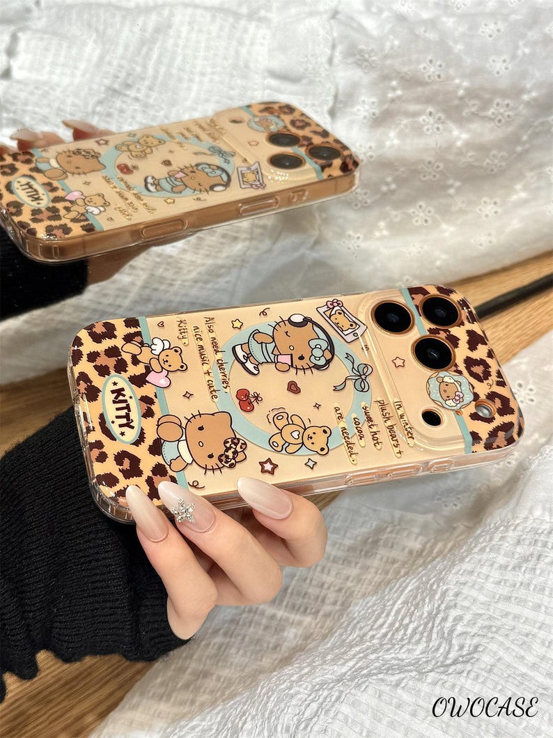 May include: Two phone cases with a tan leopard print design and illustrations of Hello Kitty characters. One case is held by a person. The cases have the word "KITTY" and text that reads "Also need cherries, nice music + cute".