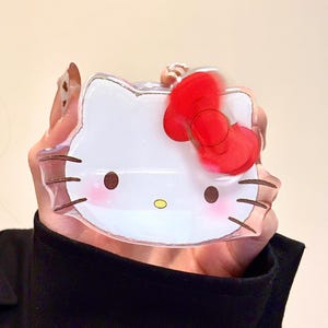 Pu&ograve; includere: Custodia per telefono trasparente e rettangolare con un design di Hello Kitty bianco e un fiocco rosso. La custodia ha baffi marroni, guance rosa e un naso giallo. La custodia &egrave; tenuta davanti a una maglietta nera.