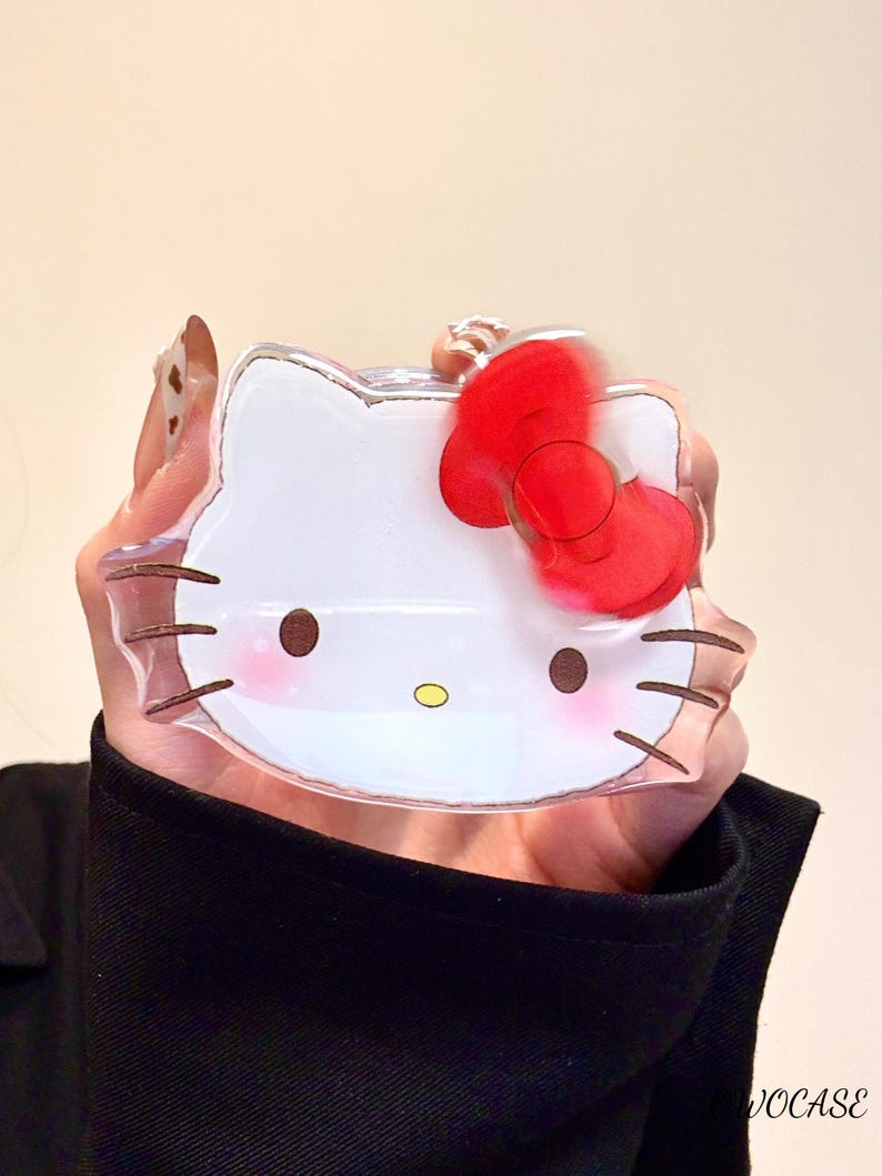 Pu&ograve; includere: Custodia per telefono trasparente e rettangolare con un design di Hello Kitty bianco e un fiocco rosso. La custodia ha baffi marroni, guance rosa e un naso giallo. La custodia &egrave; tenuta davanti a una maglietta nera.