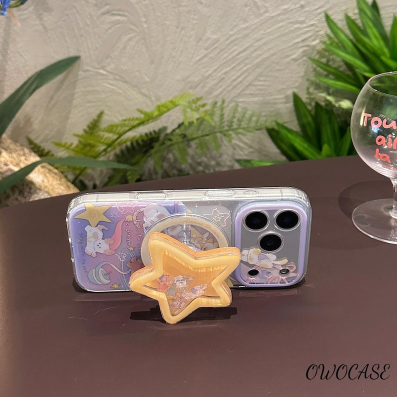 May include: Clear phone case with a whimsical design and a star-shaped phone grip. The case has a purple and white illustration with a cartoon character. The phone grip is gold and has a glittery appearance.
