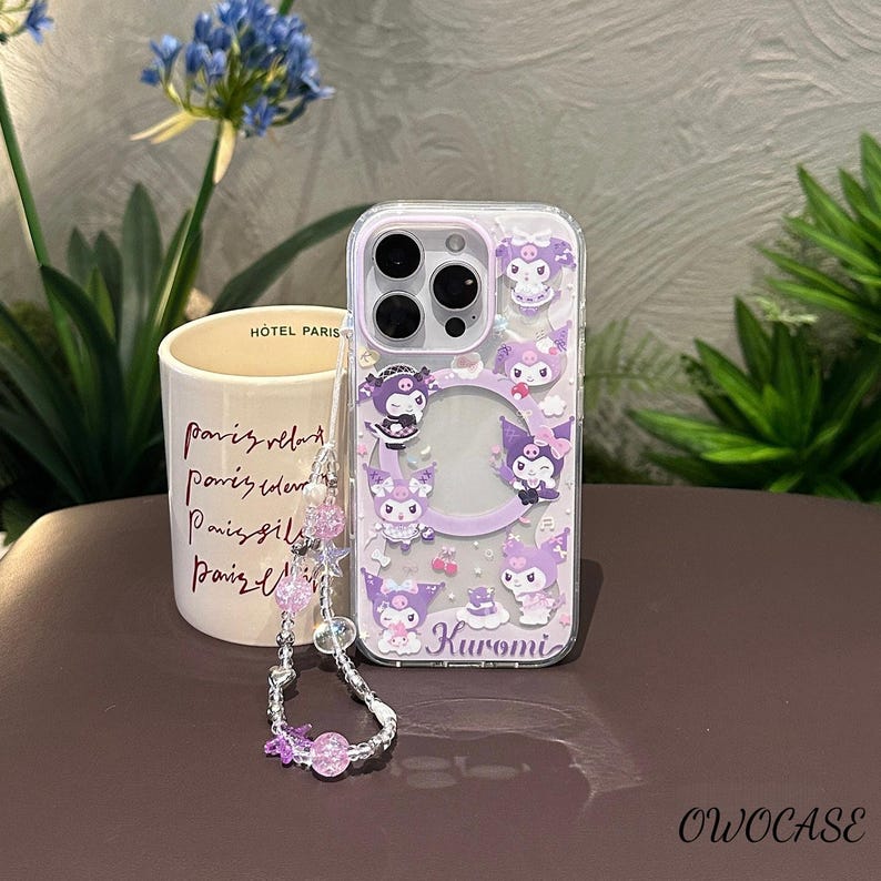 Puede incluir: Funda transparente para tel&eacute;fono con un dise&ntilde;o de Kuromi morado y blanco. La funda presenta m&uacute;ltiples ilustraciones del personaje Kuromi, un c&iacute;rculo central transparente y una correa de mu&ntilde;eca a juego con cuentas moradas. La palabra "Kuromi" est&aacute; impresa en la funda.