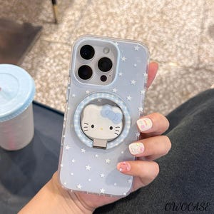 May include: Clear phone case with a light blue background and white star pattern. The case features a Hello Kitty design with a blue bow and a ring holder. The case is designed to fit a smartphone.
