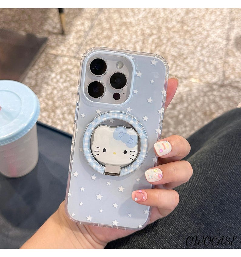 May include: Clear phone case with a light blue background and white star pattern. The case features a Hello Kitty design with a blue bow and a ring holder. The case is designed to fit a smartphone.