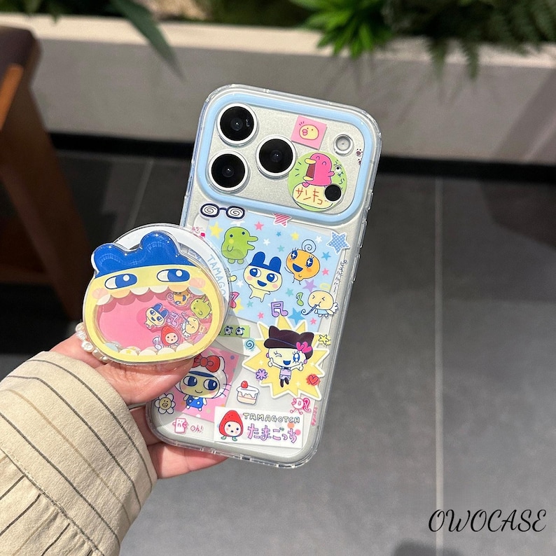 May include: Clear phone case with a blue border and colorful cartoon character stickers. A matching pop socket with a blue character design is also visible. The case and pop socket feature Japanese text and cartoon graphics.