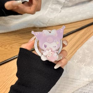 Puede incluir: Un soporte para tel&eacute;fono de color lila claro con un personaje de dibujos animados con lazo y estrellas. El soporte tiene una base blanca y un dise&ntilde;o circular. El texto "Kuromi" es visible. La marca "OWOCASE" est&aacute; en la parte inferior.
