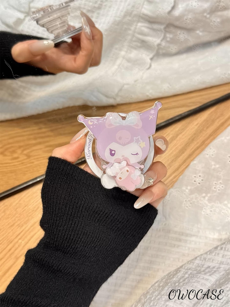 Puede incluir: Un soporte para tel&eacute;fono de color lila claro con un personaje de dibujos animados con lazo y estrellas. El soporte tiene una base blanca y un dise&ntilde;o circular. El texto "Kuromi" es visible. La marca "OWOCASE" est&aacute; en la parte inferior.