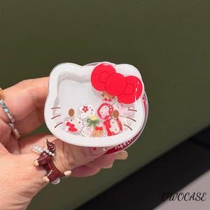 May include: A white, clear, and red Hello Kitty-shaped phone grip with a red bow. The grip has a clear center filled with small, colorful, floating charms. The text "OWOCASE" is visible on the bottom right.