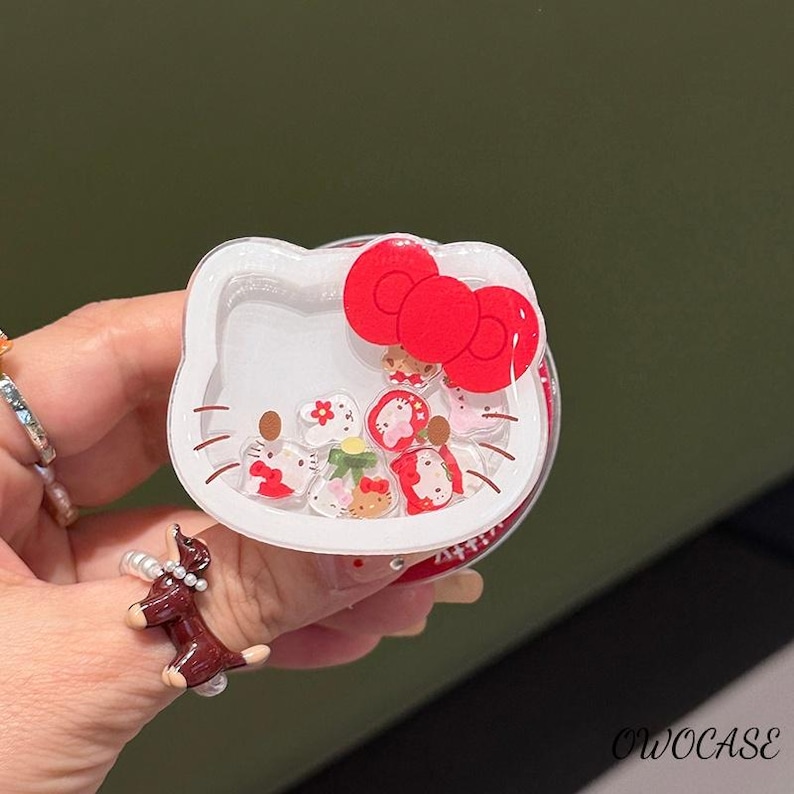 May include: A white, clear, and red Hello Kitty-shaped phone grip with a red bow. The grip has a clear center filled with small, colorful, floating charms. The text "OWOCASE" is visible on the bottom right.