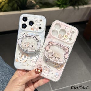 May include: Two clear phone cases with cartoon animal designs. One case is white, the other is pink. Each case has a pop-up grip with a cat ear design. The cases feature illustrations of cats, dogs, and other cute characters.
