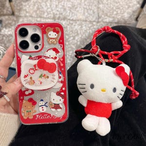 May include: A red and clear phone case with Hello Kitty graphics and a matching plush keychain. The case features a pop-up grip with a Hello Kitty design and the text "Hello Kitty". The keychain is a white plush Hello Kitty with a red bow and outfit.