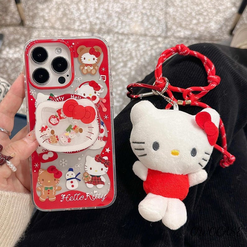 May include: A red and clear phone case with Hello Kitty graphics and a matching plush keychain. The case features a pop-up grip with a Hello Kitty design and the text "Hello Kitty". The keychain is a white plush Hello Kitty with a red bow and outfit.