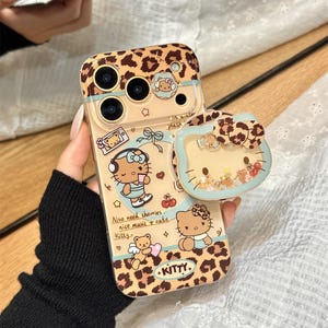 May include: A clear phone case with a leopard print design and cute cartoon illustrations. The case features the text "KITTY" and other text. A matching phone grip is attached. The case is held by a person.