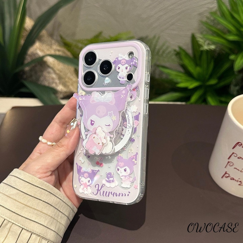 Puede incluir: Una funda transparente para tel&eacute;fono con borde lila y un agarre desplegable con un personaje de dibujos animados. La funda tiene un dise&ntilde;o con el personaje Kuromi, en tonos morados y rosas. La funda est&aacute; dise&ntilde;ada para un tel&eacute;fono con m&uacute;ltiples lentes.